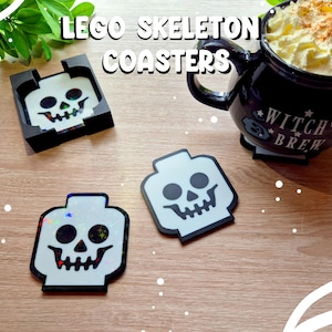 Puede incluir: Posavasos con cabeza de esqueleto estilo Lego en blanco y negro. Un posavasos está en un soporte negro. Una taza negra con las palabras "Witch's Brew" está llena de una bebida cubierta con crema batida. El texto "LEGO SKELETON COASTERS" está en la parte superior.