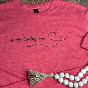 May include: Heather red t-shirt with the text "in my healing eva" and a heart design.