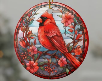 May include: A round Christmas ornament featuring a vibrant red cardinal bird perched on a branch with pink flowers. The design is framed by a stained-glass effect, with a gold string for hanging. The ornament is a festive decoration.