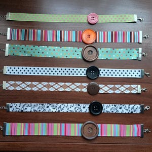 May include: A collection of seven ribbon chokers with various patterns and colors. Each choker features a large button in the center and a silver chain for adjustable sizing. Patterns include polka dots, stripes, and musical notes.