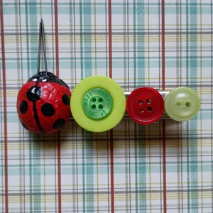 May include: A whimsical hair clip featuring a red ladybug with black spots and a row of colorful buttons. The buttons include a lime green button with text, a red button, and a light green button. The clip is set against a plaid background.