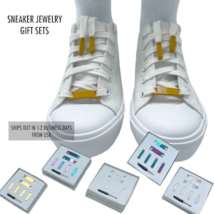 May include: Set of gold, silver, and iridescent shoe lace charms. The charms are in small boxes with a tool for attaching them to shoelaces. The charms are shaped like small rectangles.  The charms are shown on a pair of white sneakers.