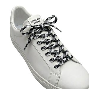 May include: A white leather sneaker featuring black and white checkered patterned shoelaces tied in a bow. A white tongue label displays the black text SHOELACE WORLD. The sneaker has a smooth finish and a standard lace-up closure system.