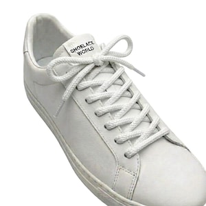 May include: White leather sneaker with white laces, featuring the text "SHOELACE WORLD" on the tongue. The shoe has a low-profile design with a white sole. The laces are neatly tied in a bow.