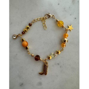 Op de afbeelding: Goudkleurige armband met amberkleurige, bruine en transparante kralen. Het bevat sterbedels, een strass-steentje en een kleine cowboylaars-hanger. De armband heeft een karabijnsluiting.