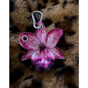 Könnte beinhalten: Ein pink-weißes Orchideenblüten-Charm mit einem silberfarbenen Verschluss. Die Blume hat detaillierte Adern und kleine, dekorative Nieten. Der Charm ist zum Anbringen an einer Tasche oder einem Schlüsselanhänger gedacht.