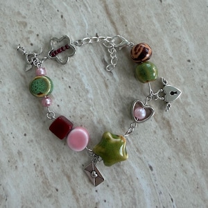 Könnte beinhalten: Ein Armband mit Perlen und verschiedenen Anhängern. Das Armband hat einen Blumen-, Herz-, Stern- und Umschlag-Anhänger. Die Perlen sind in Grün-, Rosa-, Rot- und Brauntönen gehalten.