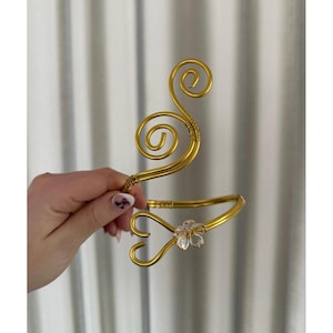Può includere: Bracciale dorato con un accento floreale. Il bracciale presenta un design a spirale con un cristallo trasparente a forma di fiore. Il bracciale è realizzato in metallo lucido color oro ed è progettato per essere indossato sulla parte superiore del braccio.