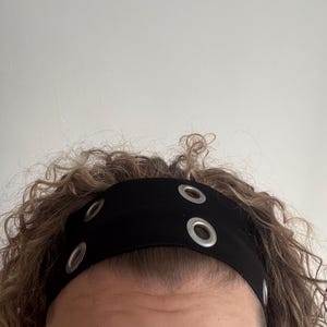 Puede incluir: Una diadema negra con tres ojales plateados. La diadema está puesta en la cabeza de una persona.