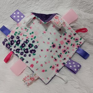 May include: A white baby blanket with a floral print and purple and pink satin ribbon tags. The blanket is soft and cuddly, perfect for a baby.