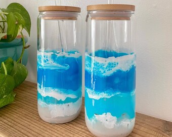 Könnte beinhalten: Zwei klare Glasbecher mit Bambusdeckeln und Glasstrohhalmen. Die Becher sind mit einer blauen und weißen Flüssigkeit gefüllt, die an Meereswellen erinnert.