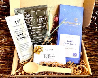 May include: A gift basket filled with brown paper shred, a wooden spoon, a small bag of coffee creamer, a bag of Vietnamese coffee, a bar of dark chocolate, and a card that says "Enjoy." The chocolate bar is blue with gold stars and the text "Moonstruck Portland Oregon Dream Dark Chocolate Isan Chocolate".