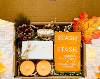 May include: A gift box filled with fall-themed items. Includes a bar of soap, two orange candles, two tea bags, a chocolate candy, and decorative elements. The box is brown with the text "Tiny Thoughts GIFT STUDIO".