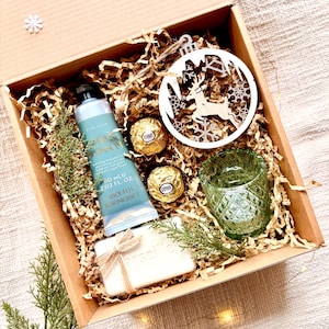 May include: A brown cardboard gift box filled with a variety of items, including a bottle of hand cream, a bar of soap, a small glass candle holder, and two chocolate truffles. The box is decorated with a wooden reindeer ornament and shredded paper.