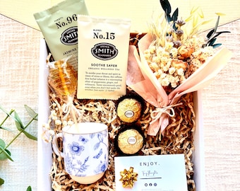 May include: A white gift box filled with a blue and white floral teacup, two chocolate truffles, a dried flower bouquet, a honey stick, and two tea bags. The tea bags are labeled "No. 96 Jasmine" and "No. 15 Soothe Sayer". The box also includes a card that says "Enjoy."