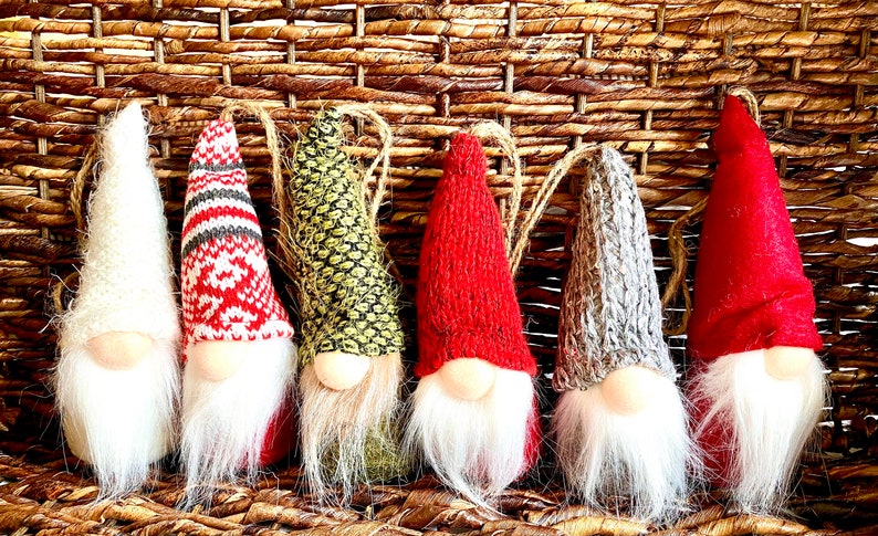 Puede incluir: Seis adornos de gnomos con sombreros de punto de diferentes colores y barbas de piel blanca. Los gnomos est&aacute;n colgados de una cesta de mimbre tejida.