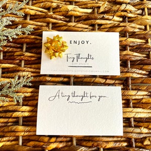 以下が含まれることがあります： 「ENJOY. Tiny Thoughts GIFT STUDIO TINY THOUGHTS.GIFTSTUDIO@GMAIL.COM」というテキストと、その上に金色のリボンが置かれた白いカード。カードの下には、「A tiny thought for you...」というテキストが書かれた別の白いカードがあります。