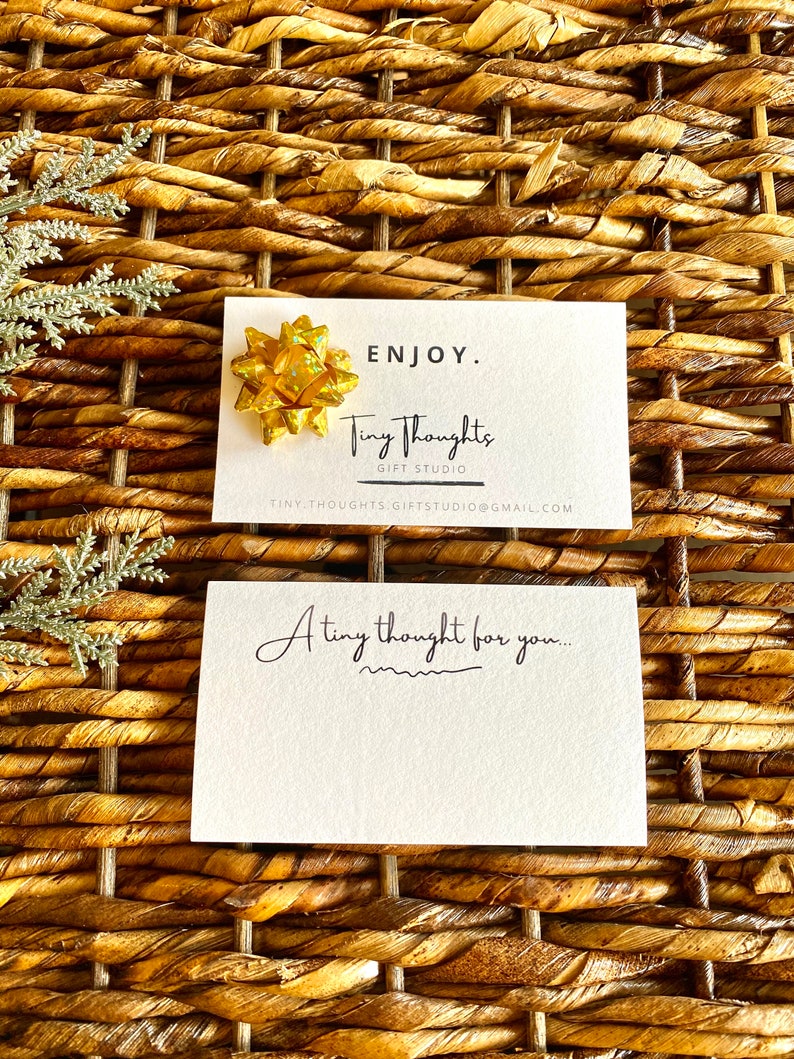 以下が含まれることがあります： 「ENJOY. Tiny Thoughts GIFT STUDIO TINY THOUGHTS.GIFTSTUDIO@GMAIL.COM」というテキストと、その上に金色のリボンが置かれた白いカード。カードの下には、「A tiny thought for you...」というテキストが書かれた別の白いカードがあります。