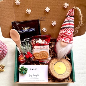 Puede incluir: Una caja de cart&oacute;n marr&oacute;n llena de un set de regalo con tem&aacute;tica navide&ntilde;a. La caja contiene una peque&ntilde;a cuchara de madera, un paquete de mezcla de chocolate caliente, un paquete de galletas de mantequilla, un peque&ntilde;o adorno de gnomo, una trufa de chocolate, una peque&ntilde;a vela en un soporte de madera y una tarjeta que dice "ENJOY. Tiny Thoughts Gift Studio".
