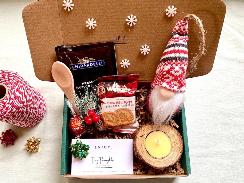 Puede incluir: Una caja de cart&oacute;n marr&oacute;n llena de un set de regalo con tem&aacute;tica navide&ntilde;a. La caja contiene una peque&ntilde;a cuchara de madera, un paquete de mezcla de chocolate caliente, un paquete de galletas de mantequilla, un peque&ntilde;o adorno de gnomo, una trufa de chocolate, una peque&ntilde;a vela en un soporte de madera y una tarjeta que dice "ENJOY. Tiny Thoughts Gift Studio".