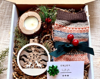May include: A Christmas gift box filled with a wooden ornament, a candle in a wooden holder, a pair of brown and orange patterned socks, and a green bow. The box is lined with brown paper and has a white card with the text "ENJOY. Tiny Thoughts" on it.