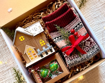 May include: A gift box filled with a small wooden house with lights, a pair of red and white patterned socks, and a box of green and white wrapped chocolates. The box is filled with brown crinkled paper and a sprig of green pine.
