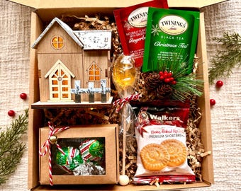 May include: A brown cardboard gift box filled with Christmas themed items. The box contains a wooden house shaped light, two tea bags, a lollipop, two chocolate candies, and a package of shortbread cookies. The tea bags are labeled "Gingerbread" and "Christmas Tea". The shortbread cookies are labeled "Walkers Home-Baked Recipe Premium Shortbread".