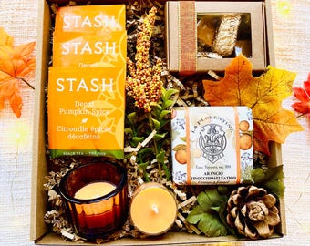 May include: A gift box filled with fall-themed items, including a box of Stash Decaf Pumpkin Spice tea, a small box of chocolates, a bar of orange-scented soap, and two candles. The box is filled with shredded paper and decorated with fall leaves and pine cones.