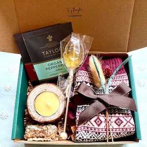 May include: A green gift box filled with a variety of items, including a pair of red and white patterned socks, a small candle in a wooden holder, a chocolate bar, a tea bag, and a lollipop. The box is tied with a brown ribbon.