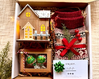 May include: A Christmas gift box with a wooden house, red and white patterned socks, a box of chocolate truffles, and a card that says "Enjoy. Tiny Thoughts".