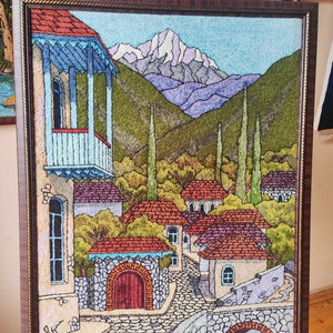 May include: A framed needlepoint artwork depicting a mountain village scene. The artwork features a mountain range in the background, a cobblestone street, and several houses with red tile roofs. The artwork is framed in a dark wood frame.