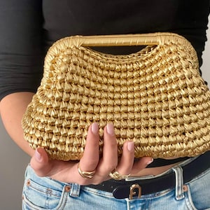 May include: A gold-colored clutch with a woven texture and a matching handle. The bag is held by a person wearing a black top and blue jeans. The clutch has a rectangular shape with rounded edges.