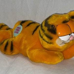 May include: Orange and black plush Garfield the cat toy laying on a white surface. The toy has a tag that says "Bored, Bored, Bored".