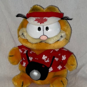 May include: Orange Garfield plush toy wearing a red and white shirt with maple leaf designs and a white headband with a red maple leaf. The plush toy is holding a black camera.