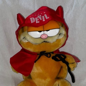 May include: A plush orange Garfield cat dressed as a devil. He is wearing a red cape with a black tie and a red headband that says "Little Devil".