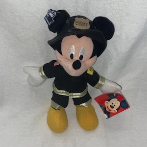 May include: A Mickey Mouse plush toy dressed as a firefighter. The plushie wears a black firefighter uniform with gold accents, a black helmet with "American Hero" on it, and yellow boots. A red tag with Mickey's face is attached.