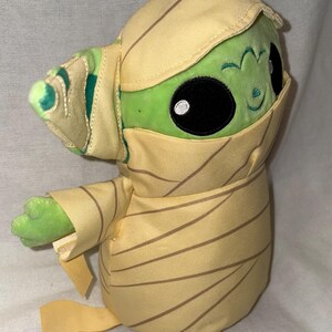 Puede incluir: Un peluche verde con grandes ojos negros y una peque&ntilde;a sonrisa, envuelto en tela beige con rayas marrones. El personaje tiene orejas grandes y lleva un gorro. El peluche es un objeto de colecci&oacute;n.