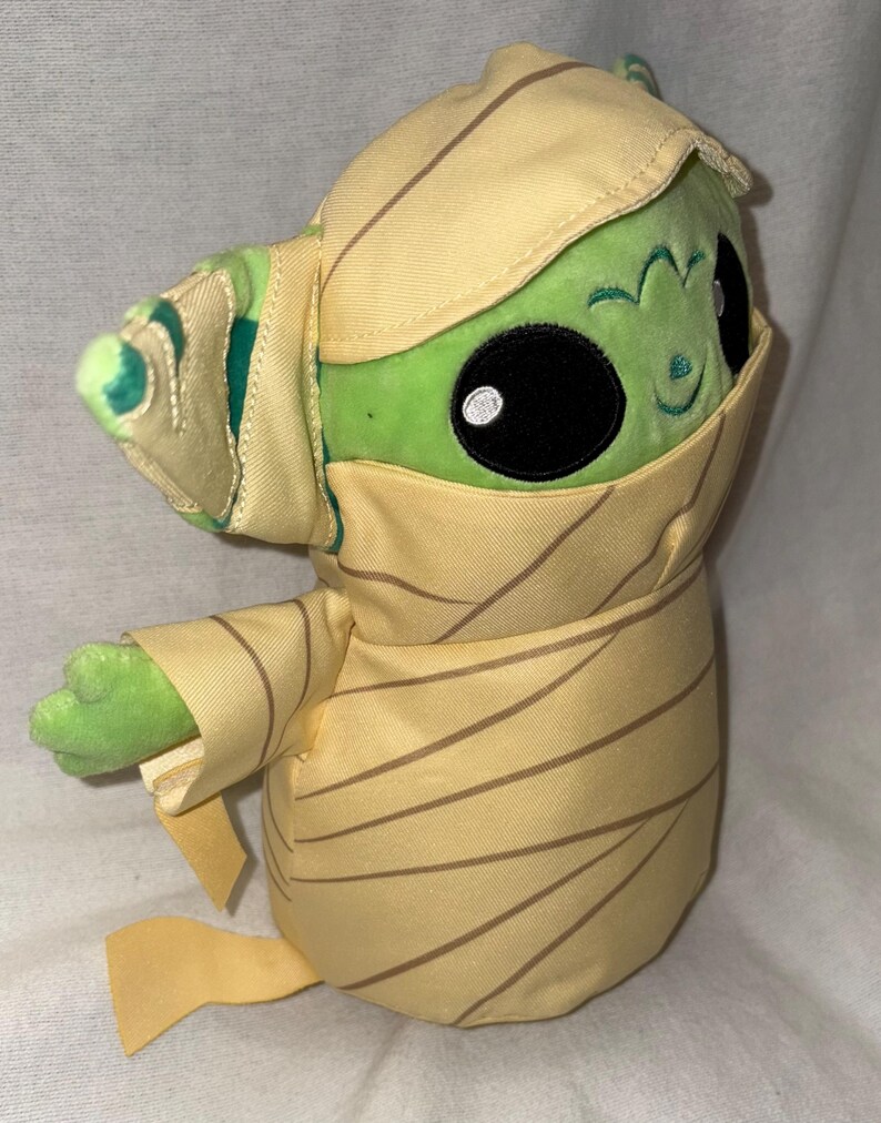 Puede incluir: Un peluche verde con grandes ojos negros y una peque&ntilde;a sonrisa, envuelto en tela beige con rayas marrones. El personaje tiene orejas grandes y lleva un gorro. El peluche es un objeto de colecci&oacute;n.