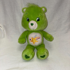 May include: A green plush Care Bear with a star and rainbow design on its belly. The bear has a heart-shaped nose and a smiling face. The plush toy is made of soft material and is a classic children's toy.