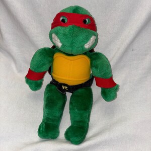 May include: A plush green turtle toy featuring a red eye mask, red wristbands, and a yellow plastron. The figure wears a black belt with a gold letter R on the buckle. The turtle has a soft texture, white teeth, and a brown shell on its back.