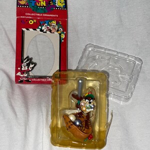 May include: A red and white box with the words "Looney Tunes" and "Collectible Ornaments" on it. The box contains a plastic ornament of Taz the Tasmanian Devil holding a baseball bat and a baseball glove with baseballs inside.