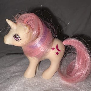 May include: A cream-colored plastic unicorn toy with a pink and purple synthetic mane and a pink tail. The figure features purple eyes and a symbol on its flank consisting of a dark pink crescent moon flanked by two small dark pink stars.
