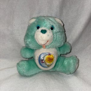 May include: A vintage teal plush Care Bear with a white belly featuring a moon and sun design. The bear has blue eyes, a green heart on its nose, and a pink tongue sticking out. It is a classic children's toy.