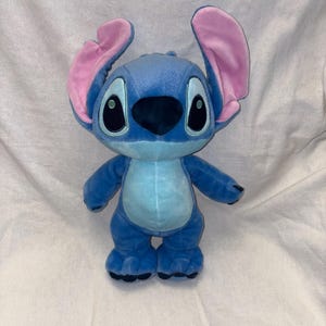 Pu&ograve; includere: Un peluche del personaggio Stitch del film d'animazione Lilo & Stitch. L'alieno blu ha grandi orecchie foderate di rosa, occhi neri e una pancia azzurra. Il giocattolo &egrave; appoggiato su una superficie bianca.