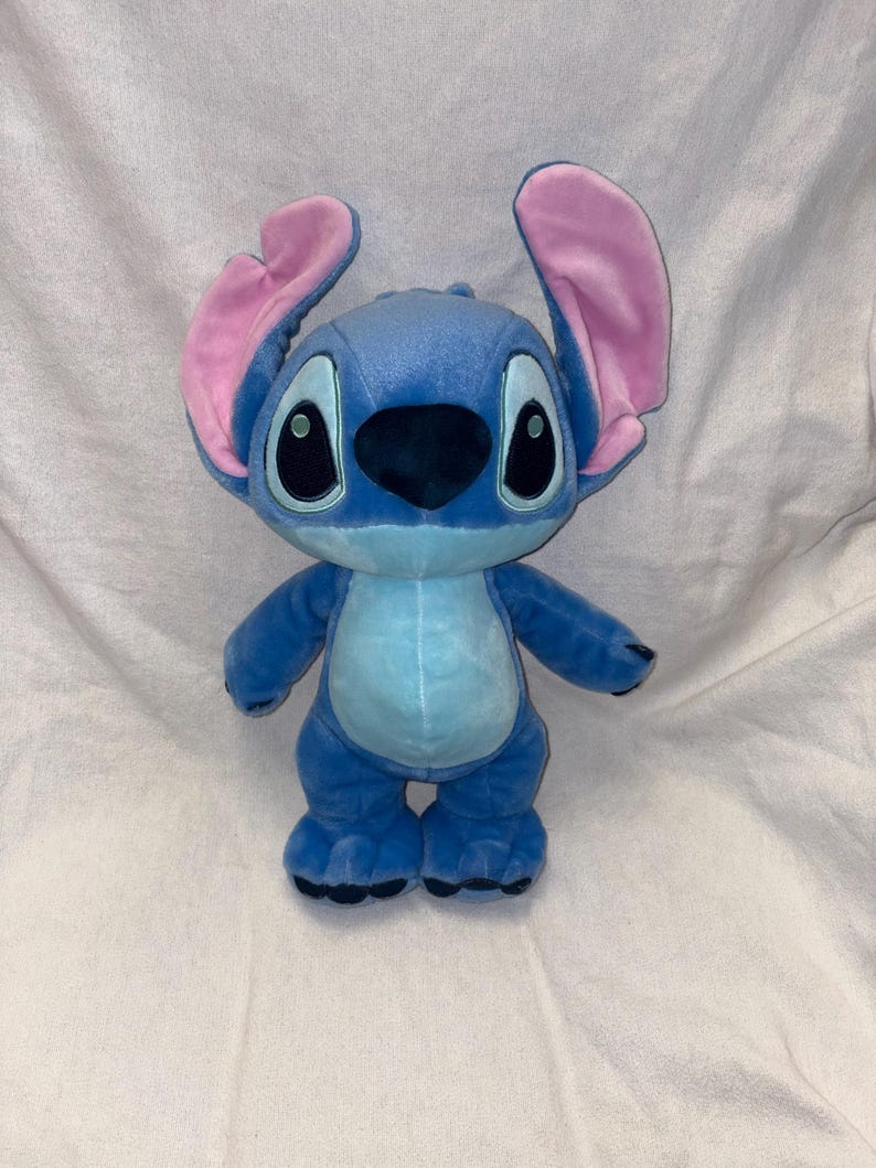 Pu&ograve; includere: Un peluche del personaggio Stitch del film d'animazione Lilo & Stitch. L'alieno blu ha grandi orecchie foderate di rosa, occhi neri e una pancia azzurra. Il giocattolo &egrave; appoggiato su una superficie bianca.