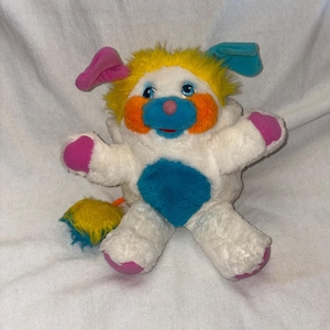 May include: A vintage plush toy in the shape of a dog-like creature. It has a white body, yellow fur, blue ears, and pink accents on the paws and ears. The face features orange cheeks, a blue nose, and a pink nose. A blue and yellow tail completes the design.
