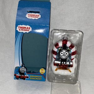 May include: Thomas & Friends Christmas ornament featuring a glass figurine of Thomas the Tank Engine wearing a candy cane hat and standing on a snowy base. The ornament is packaged in a clear plastic box with a blue and white cardboard box.