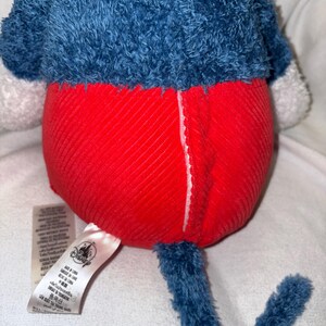 May include: A plush toy featuring a blue top and a red bottom. The toy has a blue tail and a white tag with text and a Disney logo. The toy is made of soft, textured fabric.