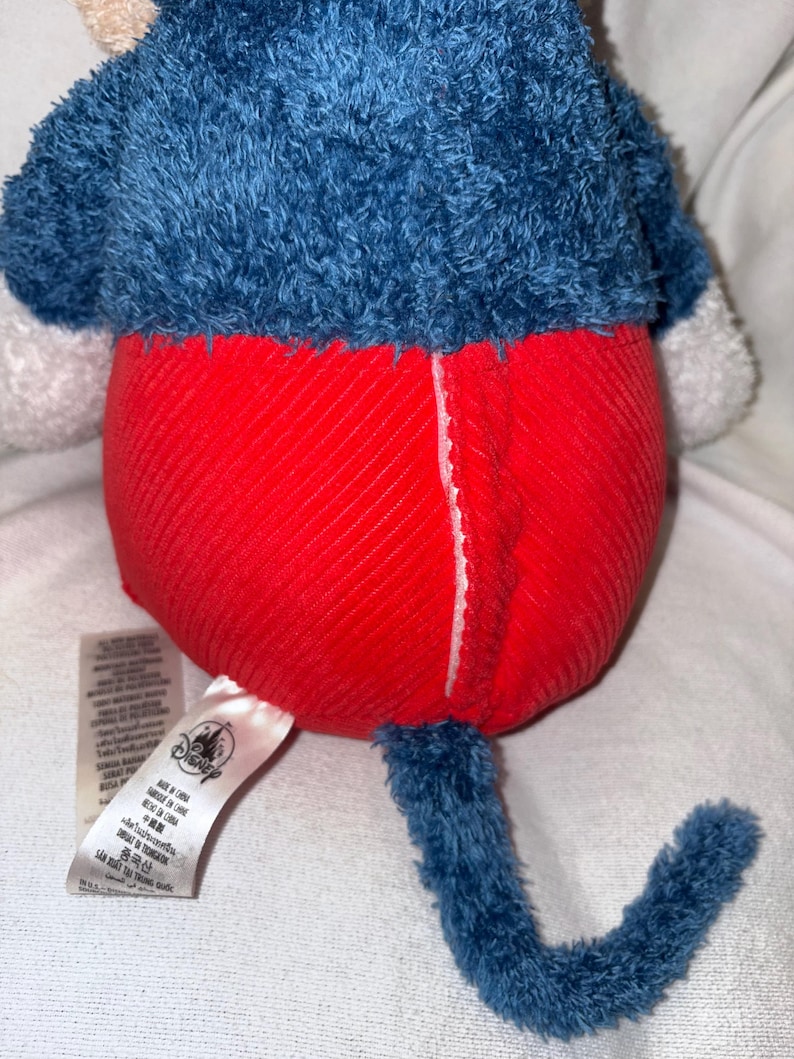 May include: A plush toy featuring a blue top and a red bottom. The toy has a blue tail and a white tag with text and a Disney logo. The toy is made of soft, textured fabric.