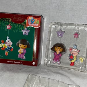 May include: A set of Dora the Explorer Christmas ornaments. The set includes Dora, Boots, and three purple stars. The ornaments are packaged in a clear plastic box with a red and white cardboard box.